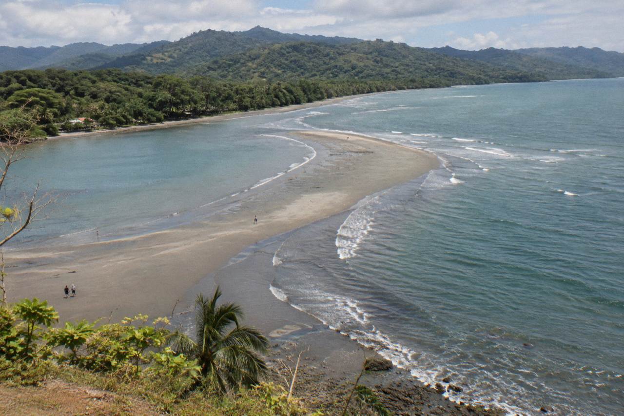 A long sandbar shaped like a whale tail curving into a calm Pacific bay, forested hills behind the beach.
