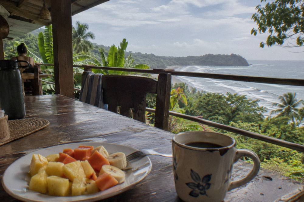 A wooden table with a plate of fresh tropical fruit and a cup of coffee beside a window overlooking the ocean
