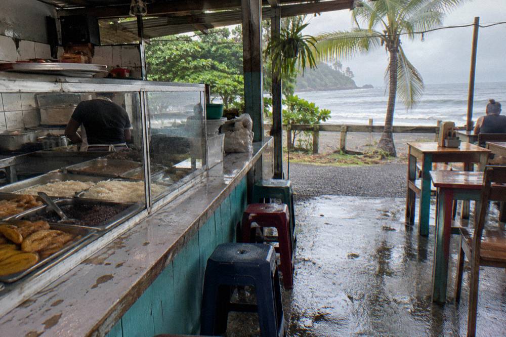 A local soda lunch counter with trays of rice, beans and plantains beside open-air seating facing the sea