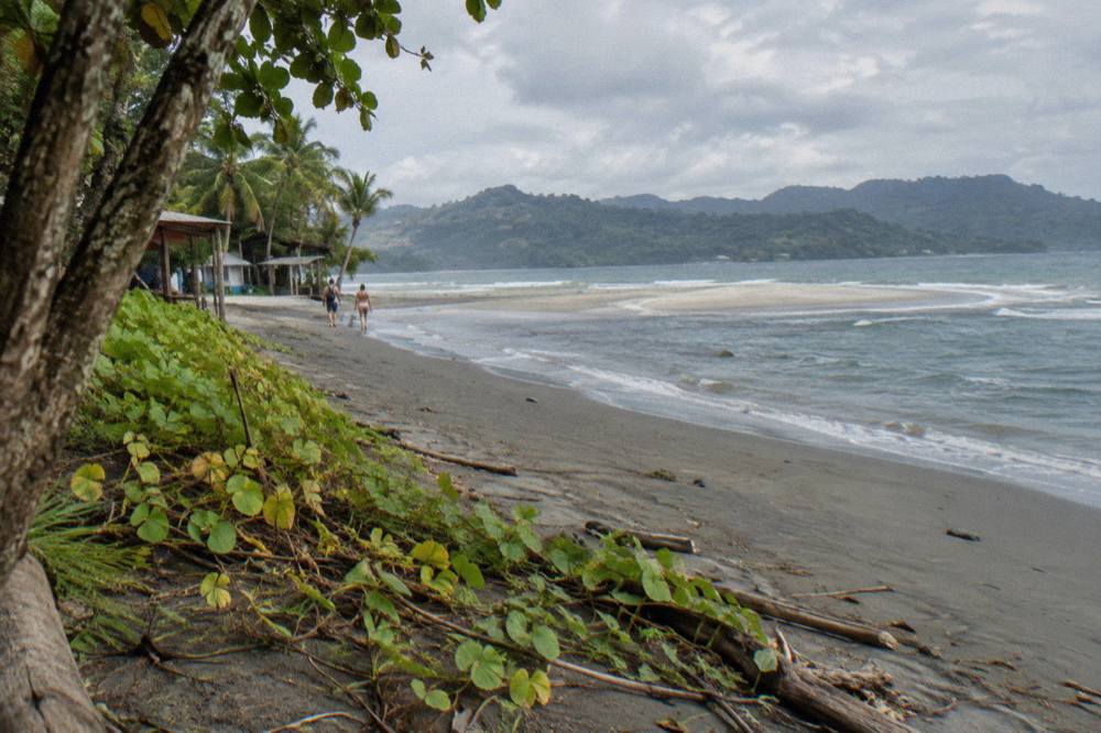 A wide empty beach at low tide with a sandbar extending into turquoise water and green hills in the distance