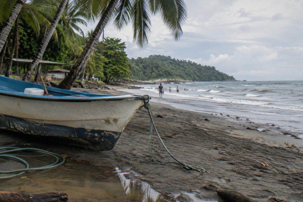 A small boat resting on a sandy beach beside palm trees with morning light over the bay