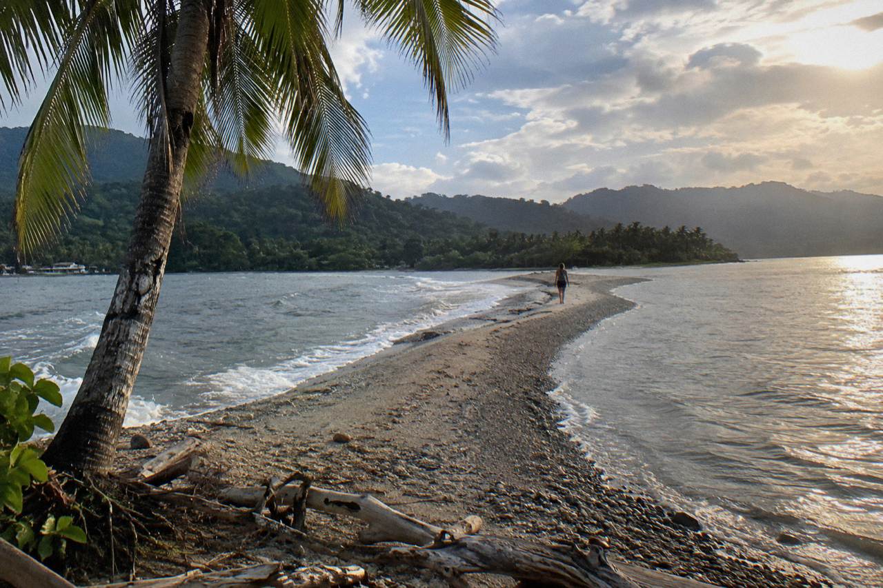 A long sandbar curving into a calm Pacific bay with forested hills behind the beach