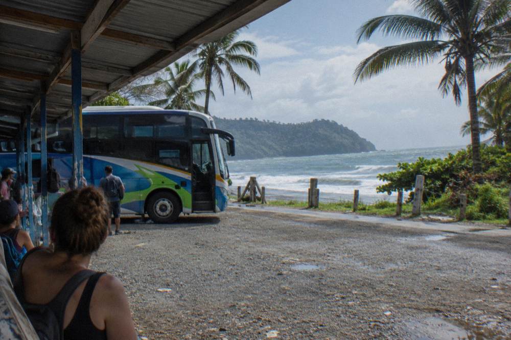 A regional bus parked at a small roadside stop with passengers waiting under a metal roof