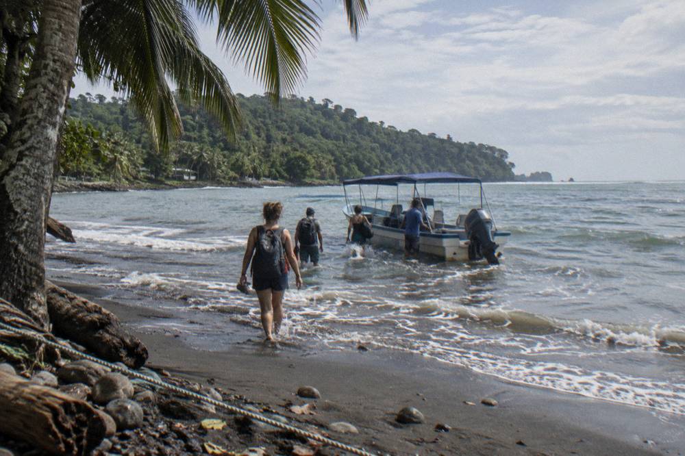 A tour boat pulled up on a dark sand beach with passengers walking toward it through shallow water