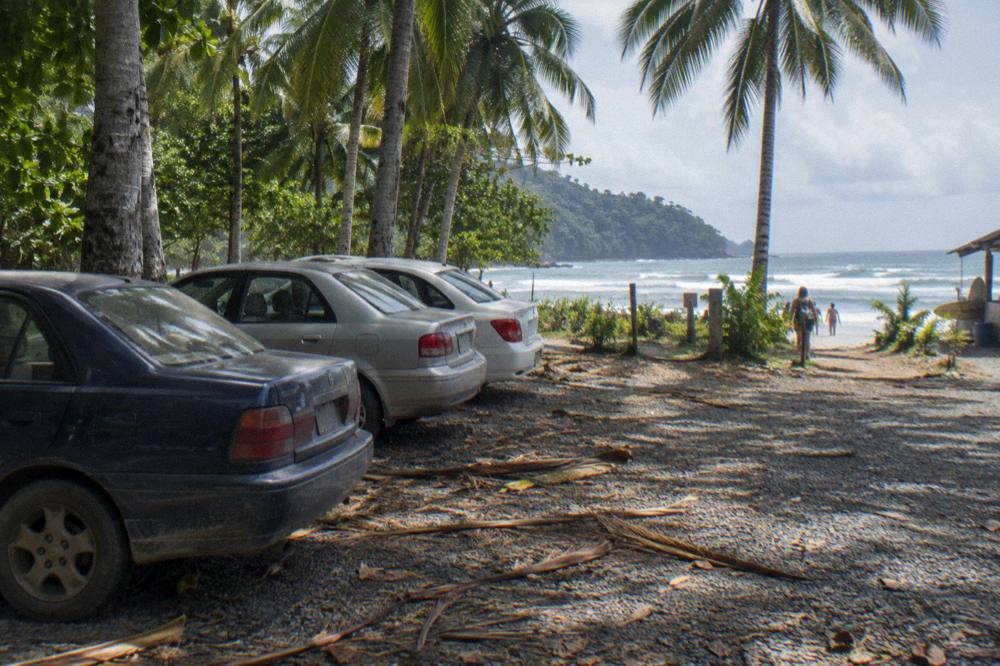 A shaded gravel parking area with a few cars beside palm trees near a beach entrance