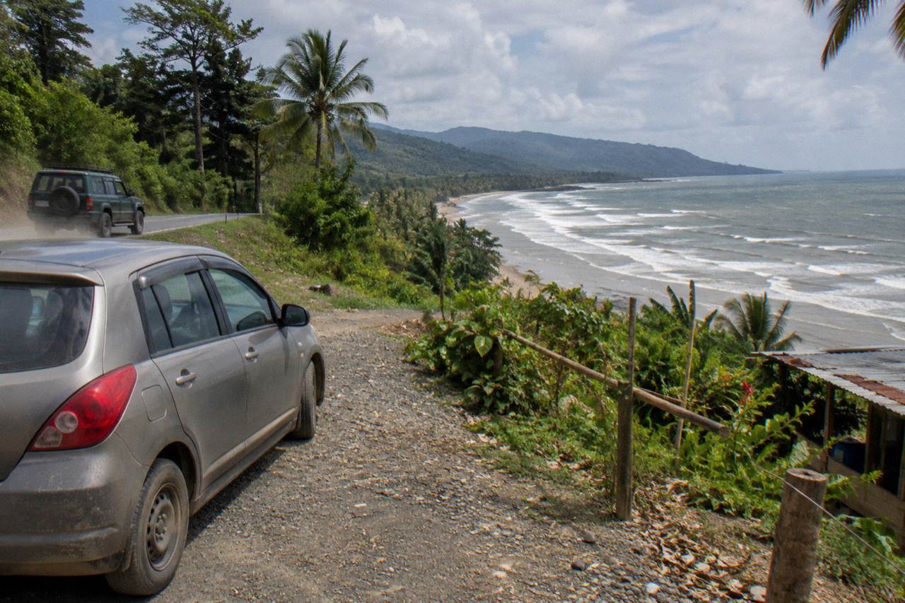A parked rental car beside the coastal road above a wide Pacific beach and green forested hills