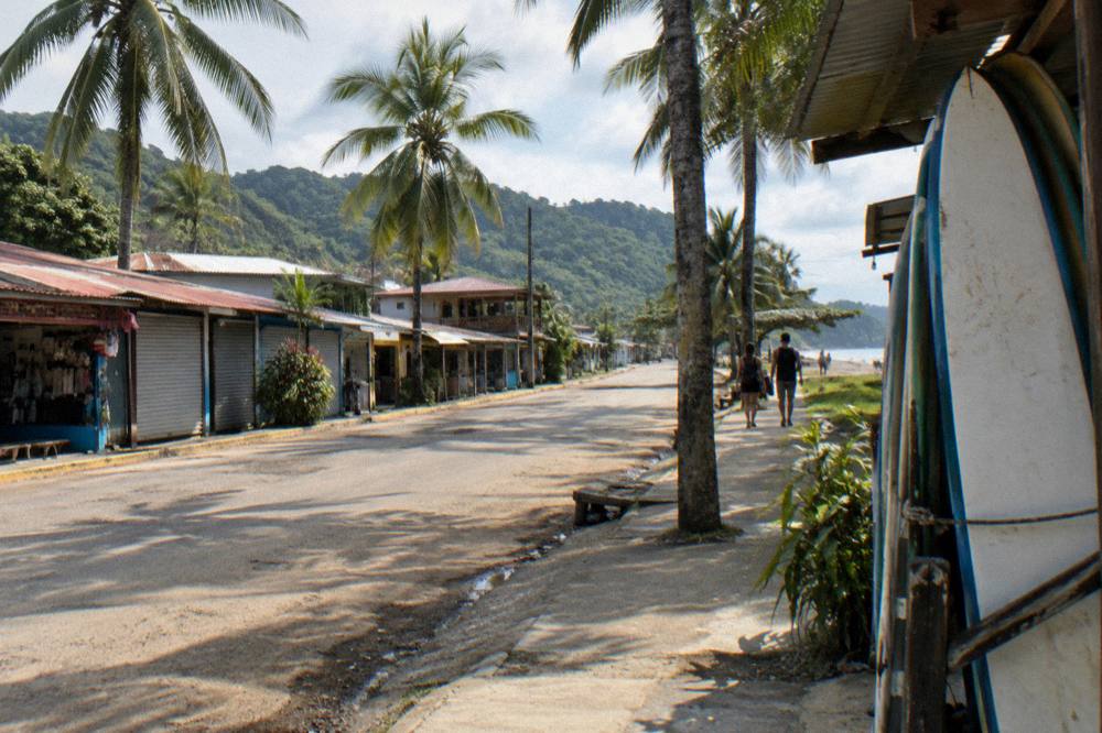 Small town street with palm trees, storefronts and hills in the background