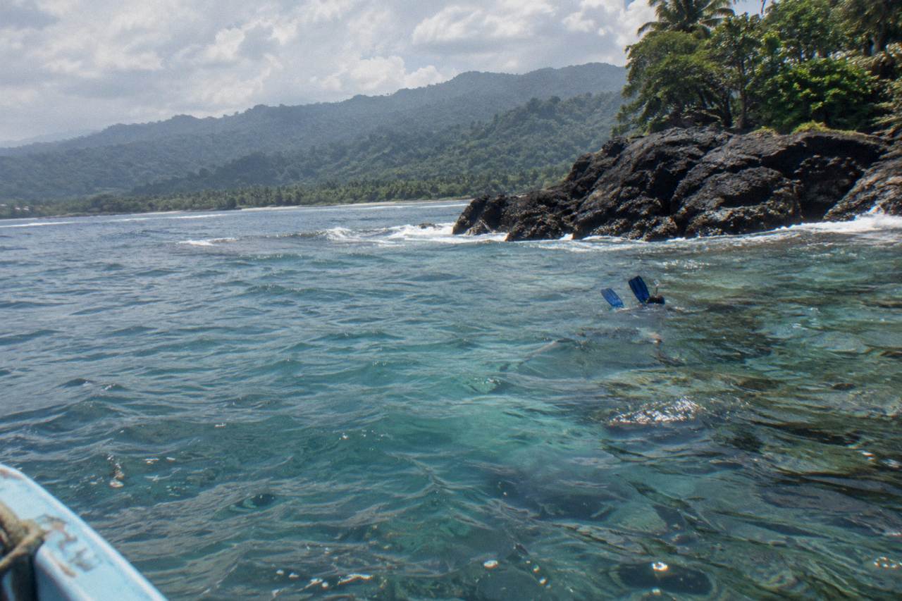 Calm turquoise water over a coral reef with rocky shoreline and forested hills behind