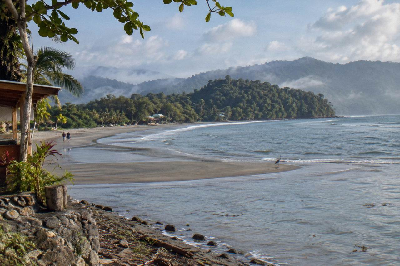 A calm Pacific bay with forested hills behind a long sandbar curving into the water