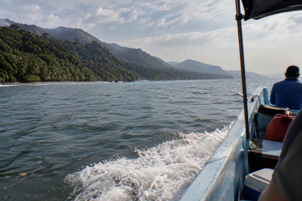 A small boat cruising close to forested green hills along a calm tropical coastline